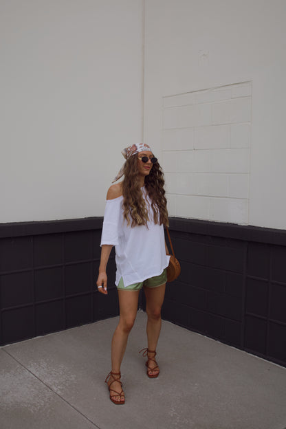 Woman in a white top and green shorts standing against a plain wall.